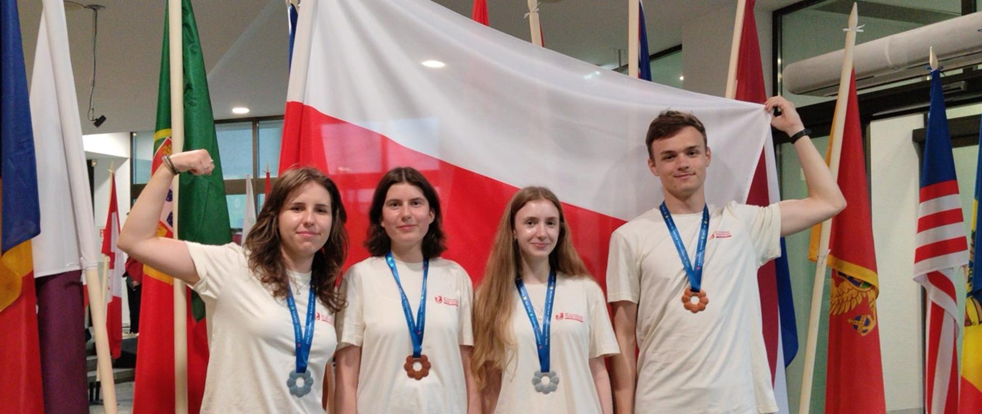 Czterch uczniów z medalami stoi w koszulkach reprezentacji Polski na tle flag państw. Uczeń z prawej strony trzyma flagę Polski.