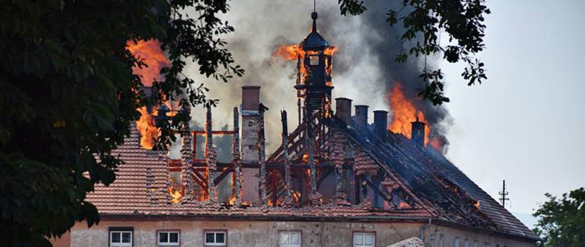 pożar budynku w Kamieńcu Ząbkowickim. Na zdjęciu widać pracę strażaków którzy gaszą pożar dachu. Na dachu budynku widać jak wydostają się płomienie. Cały dach jest obiety ogniem. Częściowo spalony. Na zdjęciu widać duża zagrożenie, ponieważ spadają dachówki.
