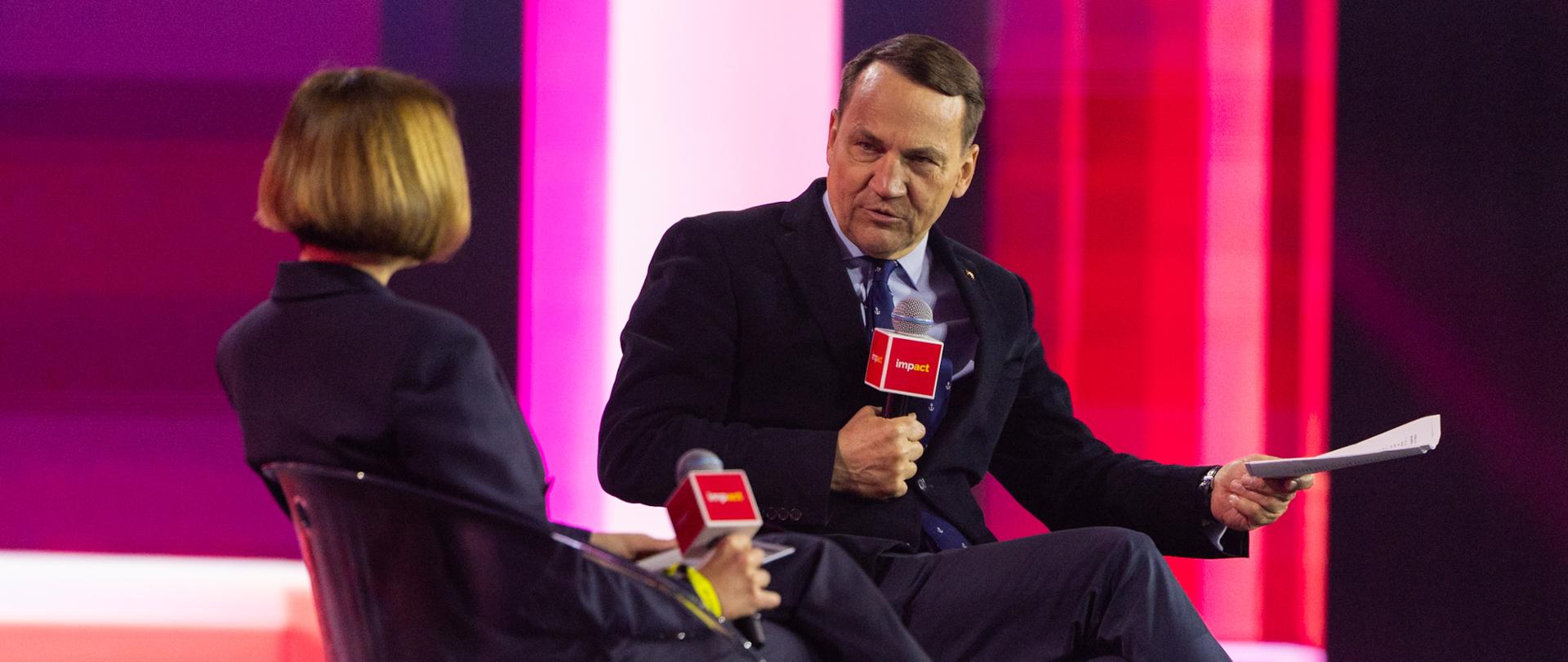 Na scenie konferencji „Impact” widać tył prowadzącej rozmowę z Wicepremierem Radosławem Sikorskim - siedzą na krzesłach podczas rozmowy panelowej. Wicepremier w ciemnym garniturze i niebieskim krawacie trzyma mikrofon oraz dokumenty, zwrócony jest twarzą do aparatu.