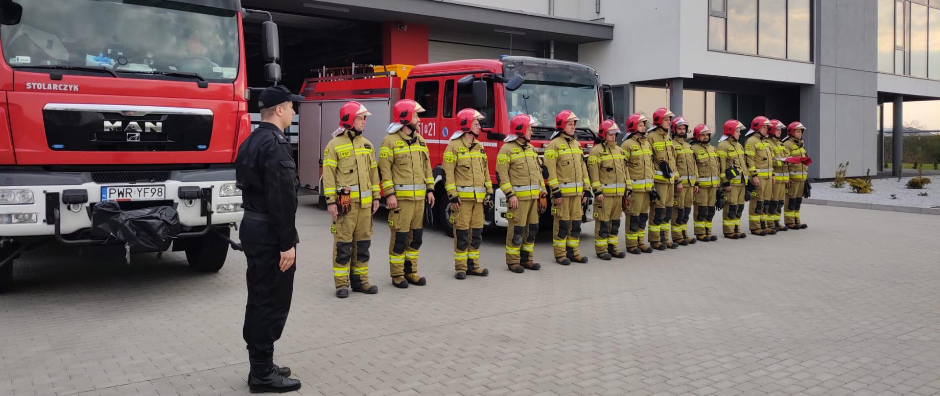 Zdjęcie przedstawia piętnastu strażaków stojących przed budynkiem komendy. Za nimi dwa wozy 