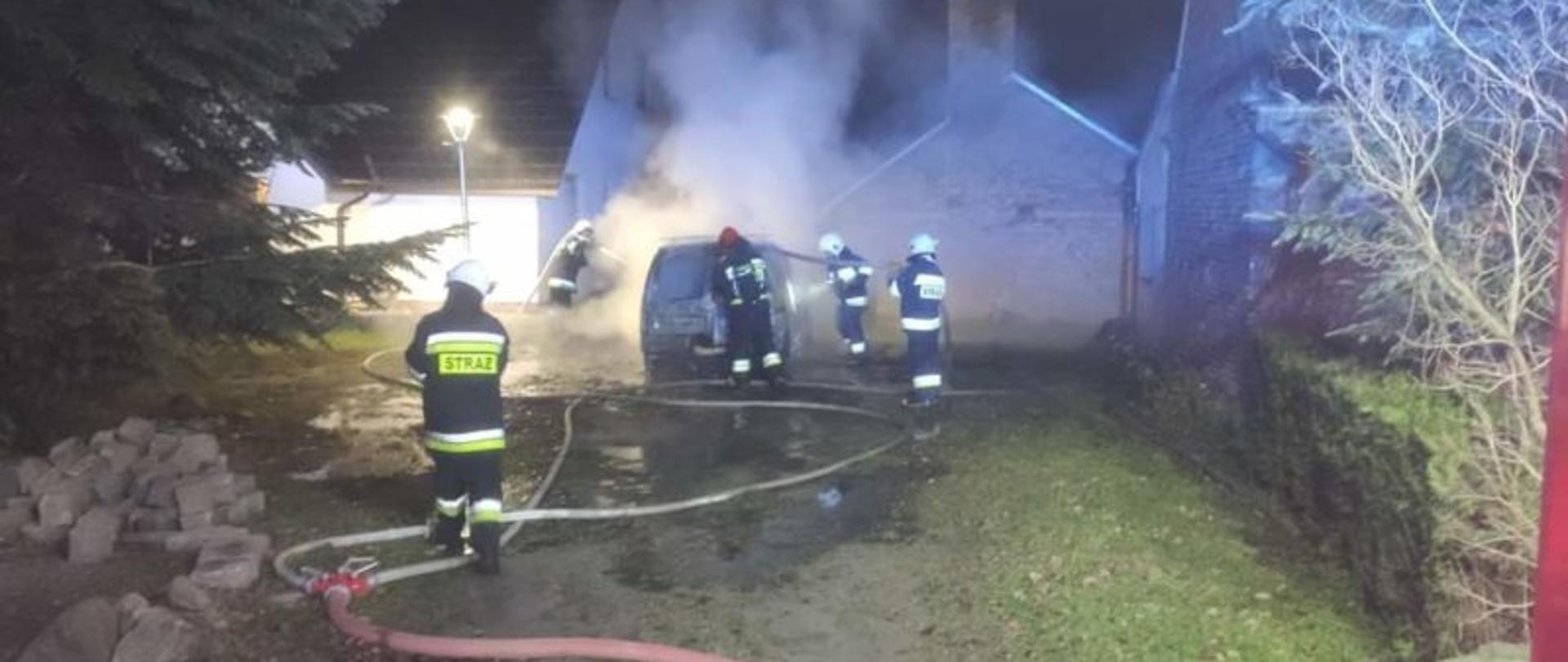 Strażacy gaszą samochód osobowy. Widoczne linie wężowe, okoliczne zabudowania. Dużo dymu.