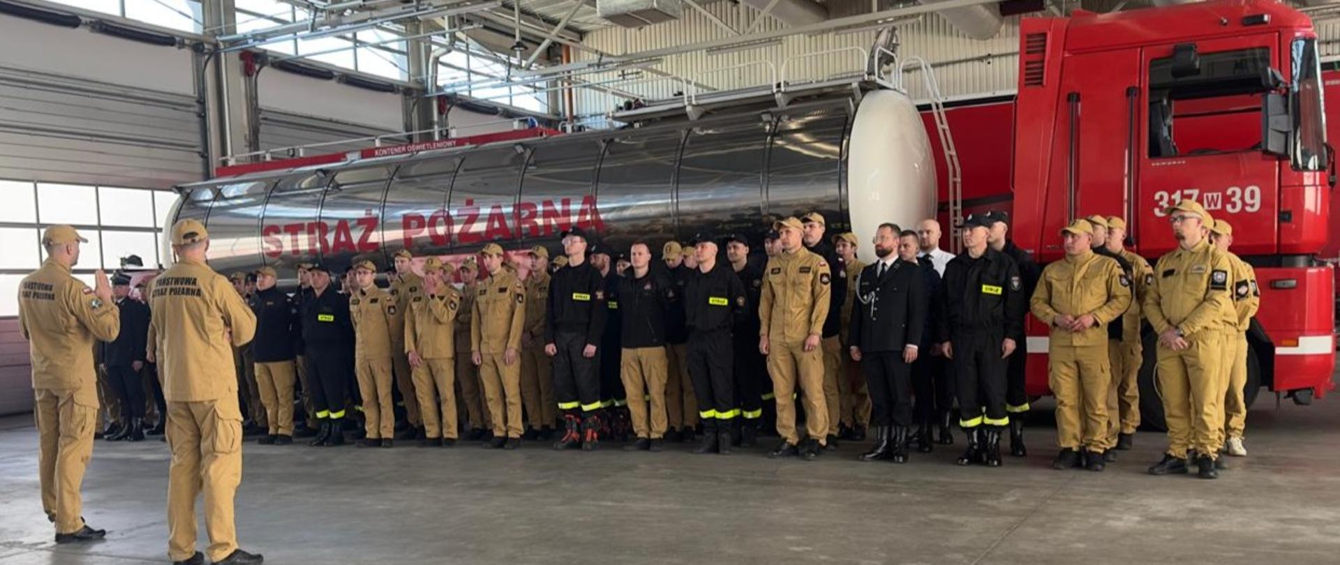 2 instruktorów widocznych tyłem tyłem. Ćwiczące poczty sztandarowe stoją na tle samochodu cysterny. 