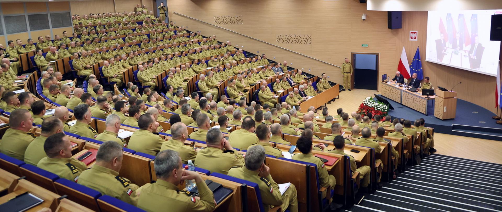 Widok ogólny na aule podczas narady służbowej. Strażacy w mundurach służbowych w kolorze piaskowym siedzą w auli Centralnej Szkoły Państwowej Straży Pożarnej w Częstochowie