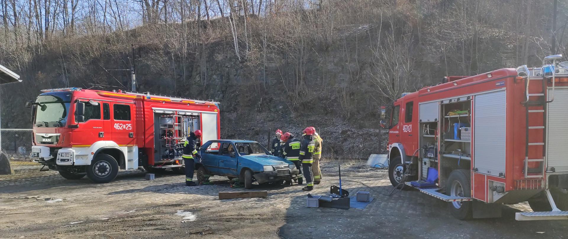 Pięciu strażaków w ubraniu specjalnym stojący przy zniszczonym samochodzie osobowym koloru niebieskiego. Po prawej i lewej stronie dwa samochody ratowniczo-gaśnicze