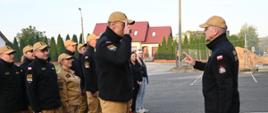 Strażacy stoją ustawieni w dwóch rzędach. Przed nimi na środku stoi dwóch strażaków oddają sobie honor przez salutowanie. W tle zabudowa osiedlowa i plac budowy.