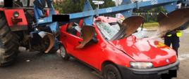 Widać mocno uszkodzony czerwony samochodu od przodu, w szybę przednią są wbite brony