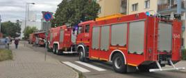 Na zdjęciu widnieją wozy strażackie podczas akcji gaszenia pożaru mieszkania w budynku wielorodzinnym. Dzień, jasno, bez opadów. 