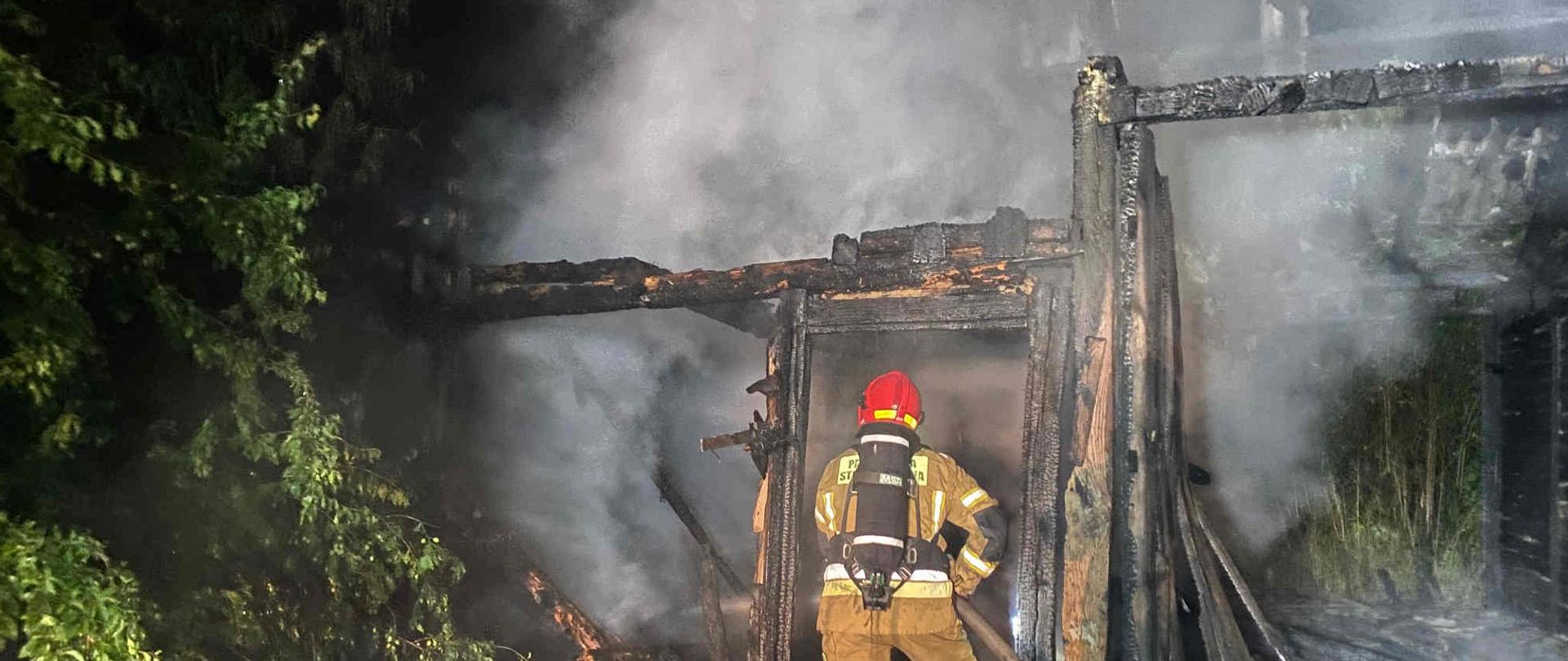 Zdjęcie zrobione w nocy. Na zdjęciu widać palącą się stodołę, dym oraz strażaka ubranego w sprzęt ochrony układu oddechowego trzymającego wąż pożarniczy, podającego wodę na palące się elementy konstrukcyjne budynku gospodarczego. 