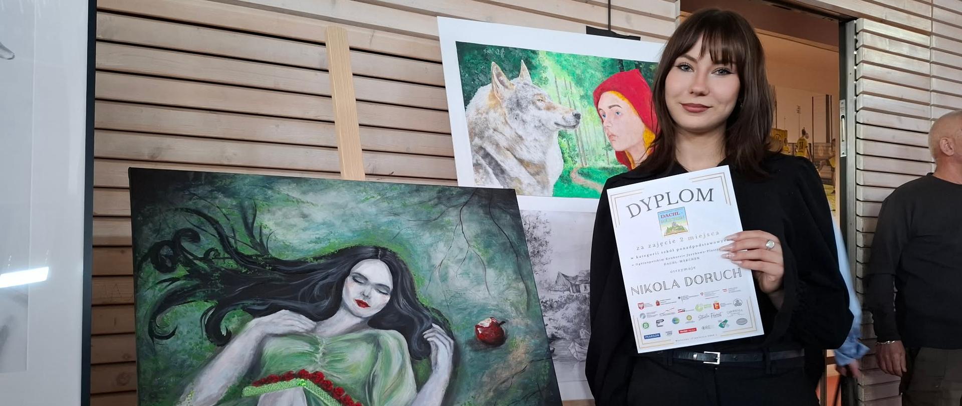 Dziewczyna z dyplomem w ręce, po jej lewej stronie obraz na sztaludze przedstawiający Królewnę Śnieżkę, która zasnęła po ugryzieniu zatrutego jabłka. z tyłu na szlugach prace ilustrujące inne bajki min. Czerwonego Kapturka