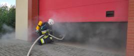 Zdjęcie przedstawia strażaka, który asekuruję pracę prądownika i trzyma linię gaśniczą. Strażacy pracują w zadymieniu i mają na sobie założone ubrania specjalne, hełmy oraz aparaty powietrzne podłączone do masek na twarzy. Na zdjęciu widać część bramy garażowej, przez którą wchodzą strażacy. 