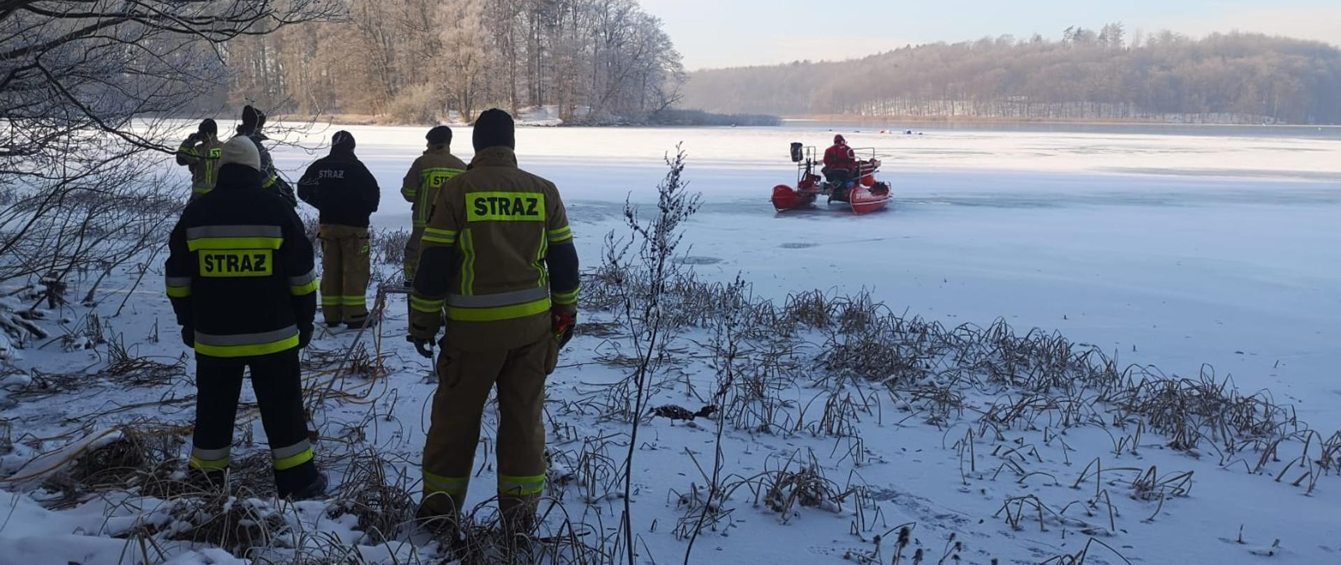 Sanie typu woda-lód w trakcie poszukiwań na jeziorze. Na brzegu znajdują obserwujący działania strażacy.