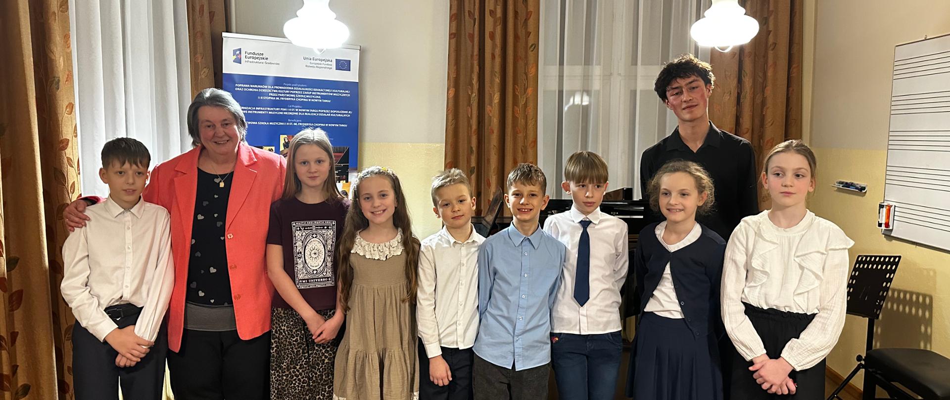 Zdjęcie przedstawia grupę dziesięciu osób – dwoje dorosłych i ośmioro dzieci – pozujących w pomieszczeniu, które wygląda na salę muzyczną lub lekcyjną. Po lewej stronie stoi uśmiechnięta, starsza kobieta w koralowej marynarce, obejmująca ramieniem chłopca. W centrum i po bokach znajduje się ośmioro dzieci (pięciu chłopców i trzy dziewczynki) w wieku szkolnym. Są ubrane odświętnie: chłopcy w koszulach (białych i błękitnej), niektóre z krawatami, a dziewczynki w sukienkach lub spódnicach i eleganckich bluzkach. Z tyłu, po prawej stronie, stoi wysoki młody mężczyzna w czarnej koszuli. Instrumenty i sprzęt: W tle, za grupą, widoczny jest fragment czarnego fortepianu. Po prawej stronie stoi pulpit na nuty oraz tablica z narysowaną pięciolinią. Wystrój: Okna zasłonięte są długimi, wzorzystymi zasłonami w odcieniach brązu i złota. Z sufitu zwisają dwie białe, kuliste lampy. Informacje dodatkowe: Za grupą stoi niebieski baner z logo Unii Europejskiej oraz napisami w języku polskim, co sugeruje, że zdjęcie zostało zrobione podczas oficjalnego wydarzenia, np. koncertu lub zakończenia warsztatów muzycznych.