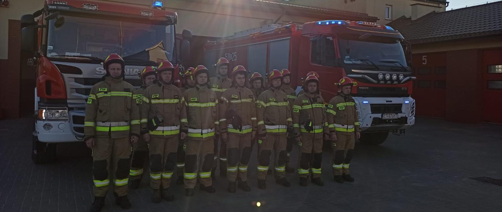 Zdjęcie przedstawia trzynastu strażaków znajdujących się na placu Komendy Powiatowej Państwowej Straży Pożarnej w Rypinie, podczas zmiany służby. Funkcjonariusze ustawieni są w dwuszeregu i stoją na baczność. W ten sposób poprzez minutę ciszy oddają cześć dla bohaterskich ratowników Ukrainy. Za strażakami znajdują się dwa samochody gaśnicze z włączonym oświetleniem alarmowym. W tle widać budynek Komendy Powiatowej Państwowej Straży Pożarnej w Rypinie.