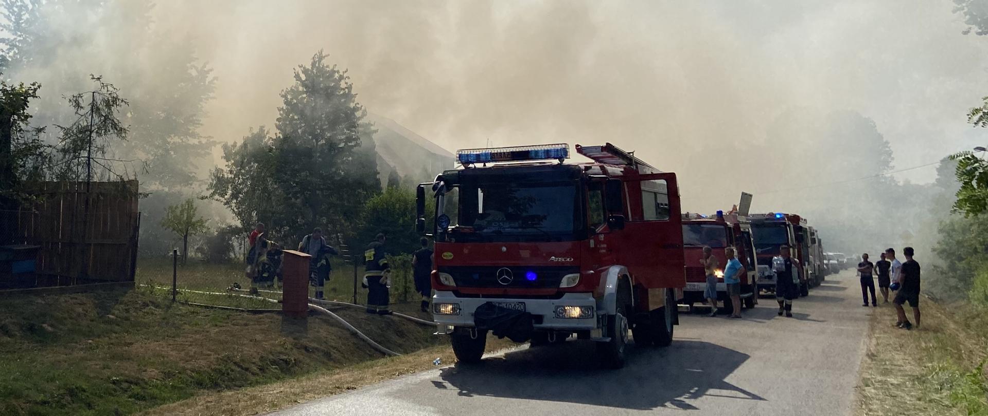 Na zdjęciu widzimy samochody strażackie stojące na poboczy drogi, w tle zadymienie.