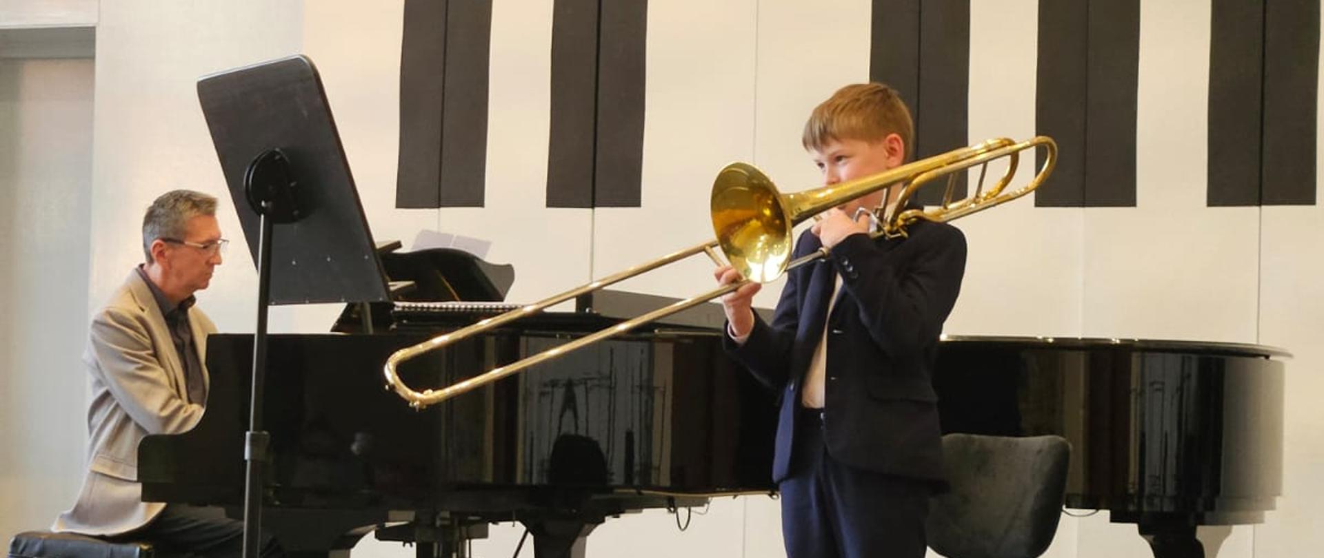 Chłopiec gra na puzonie, mężczyzna akompaniuje na fortepianie.
