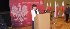 Dzień Strażaka w Rawiczu. Sala widowiskowa w domu kultury. Na tle ścianki reklamowej, przy mównicy stoi pani minister, która wygłasza okolicznościowe przemówienie do obecnych na uroczystości. Z prawej strony stoi stojak z flagami.