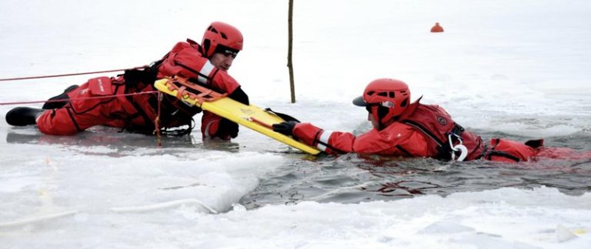 Na zdjęciu ratownik leżących na lodzie wyposażonych w deskę ratowniczą pomaga wydostać się z przerębli drugiemu ratownikowi, który odgrywa rolę osoby poszkodowanej pod którą załamał się lód.