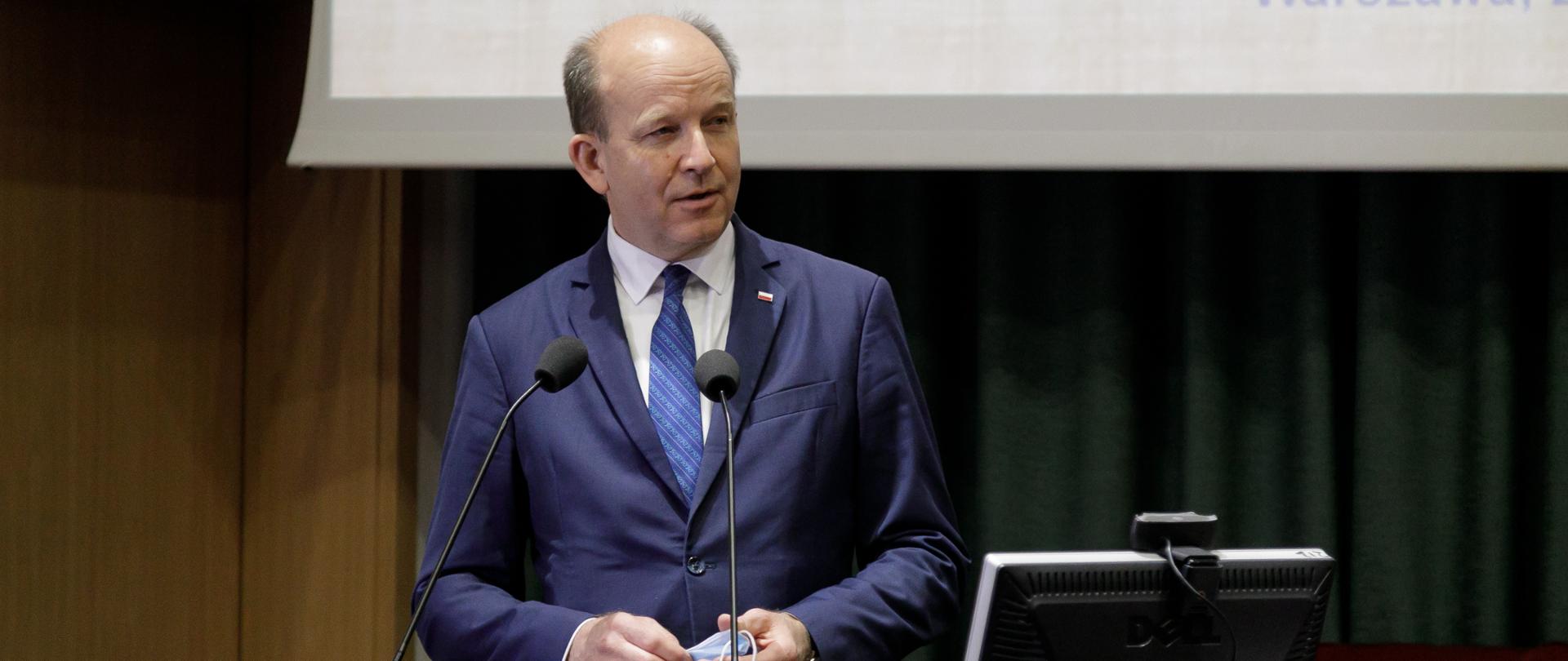 Wojewoda Mazowiecki ubrany w granatowy garnitur stojący za brązową mównicą 