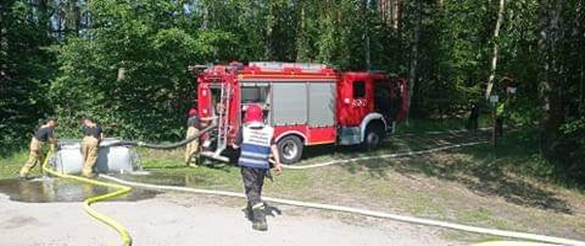 Zdjęcie przedstawia wóz straży pożarnej w lesie w czasie ćwiczeń. Strażacy pompujaąwode do rozkładanego zbiornika z wodą.