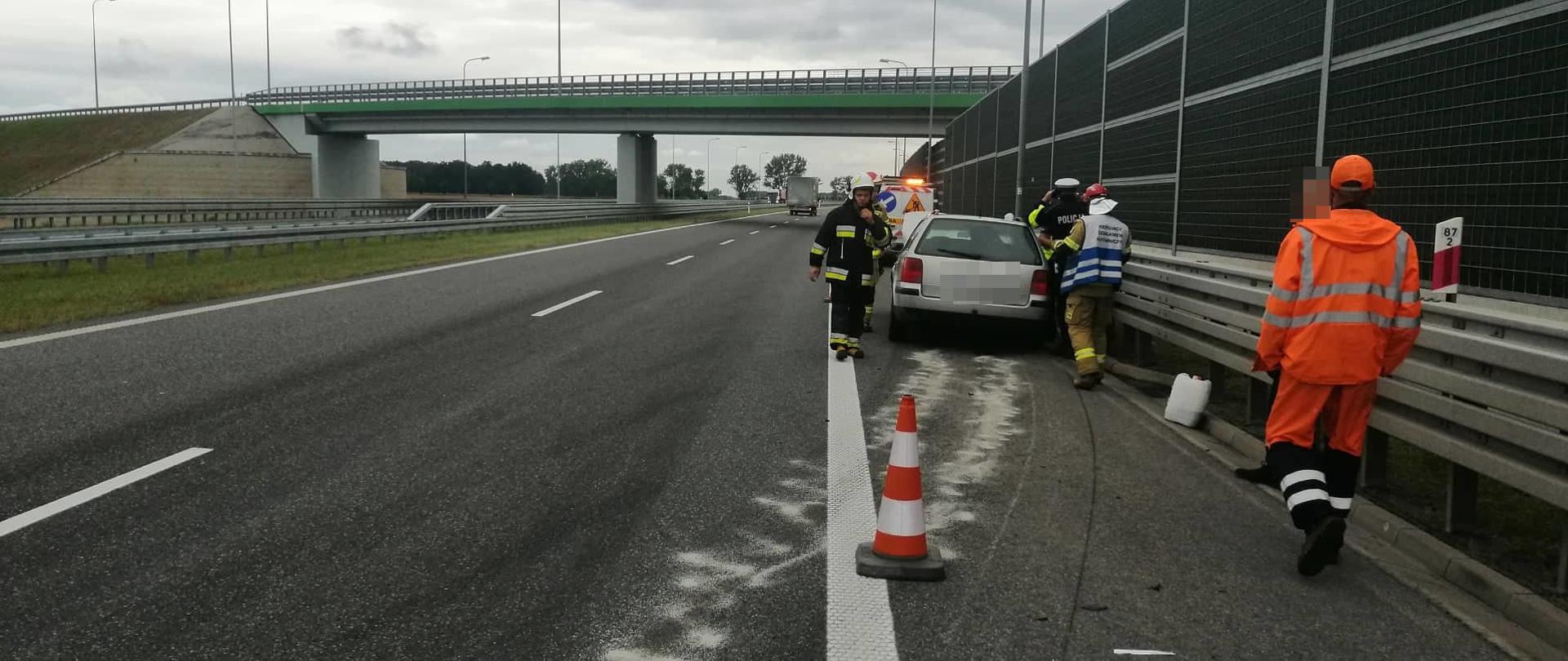 Kolizja na s5. Jezdnia drogi ekspresowej, pikietaż 87,2. Na pasie awaryjnym stoi samochód osobowy, który uderzył w barierę energochłonna. Przed nim pojazd służby drogowej. Obok znajdują się strażacy, policjanci oraz pracownik służby drogowej. W tle wiadukt.