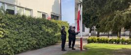 Na zdjęciu widać poczet flagowy przy maszcie - trzech funkcjonariuszy w mundurach wyjściowych. Dwóch z nich oddaje honor, środkowy zaś dokonuje podniesienia flagi państwowej.