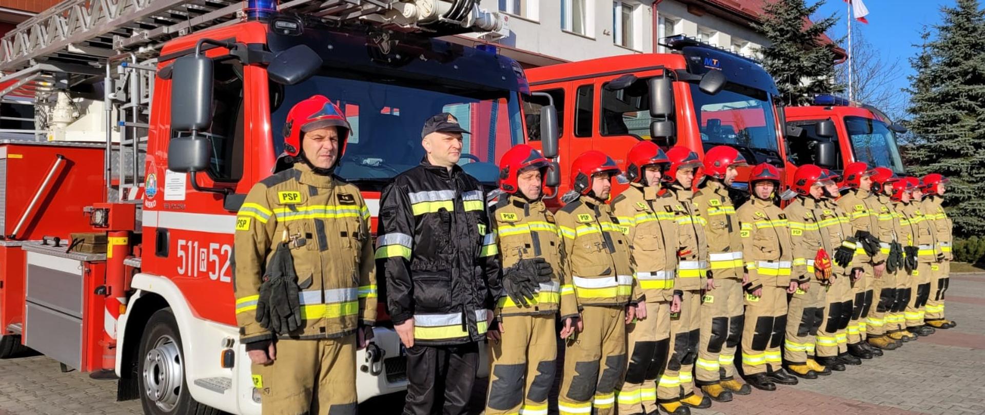 Zdjęcie zrobione przed Jednostką Ratowniczo-Gaśniczą Państwowej Straży Pożarnej. Strażacy w umundurowaniu uczestniczą w uroczystej zbiórce. Ustawieni są w szeregu. Za nimi stoją samochody pożarnicze.