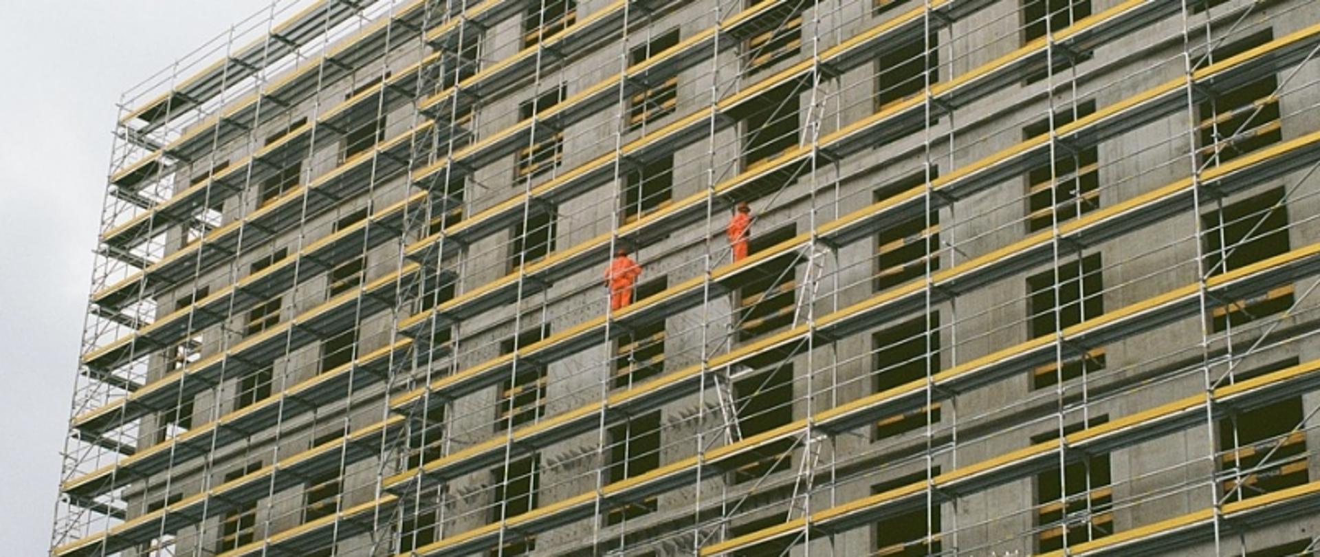 Betonowy budynek w budowie z rusztowaniem i dwoma pracownikami w pomarańczowych strojach ochronnych.