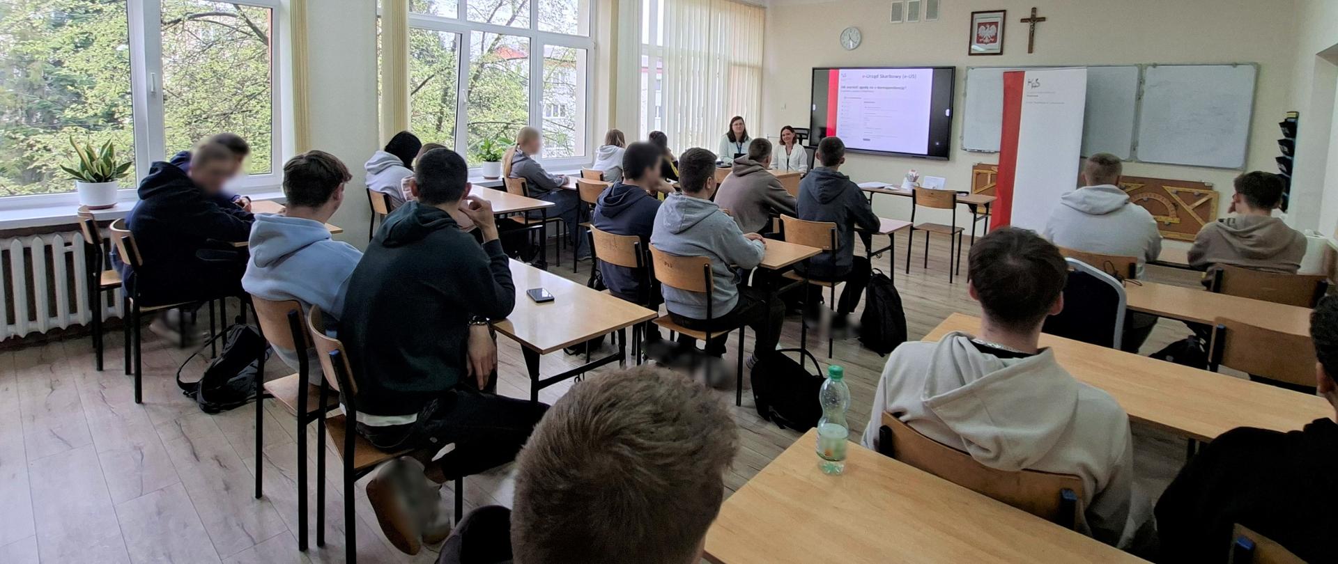 Młodzież siedzi w sali lekcyjnej Zespołu Szkół. Przed nimi są ekspertki KAS. Na tablicy wyświetlana jest prezentacja. 