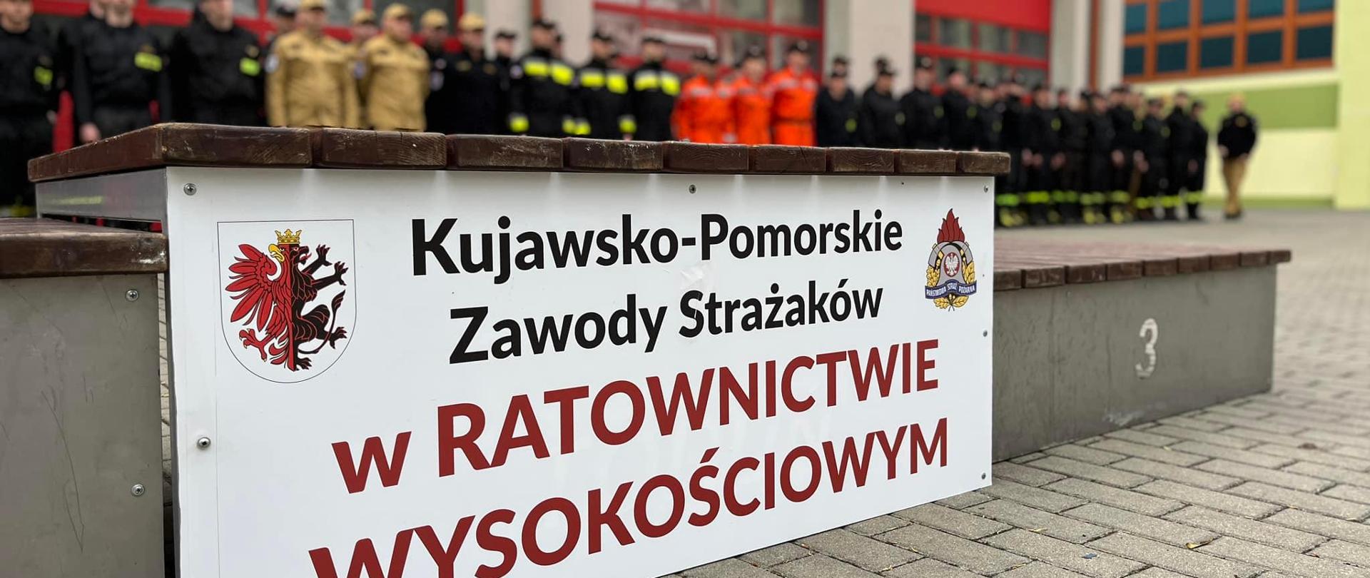 Plansza przytwierdzona do podium. Na niej napis z nazwą zawodów. W tle zarys kilkudziesięciu zawodników.