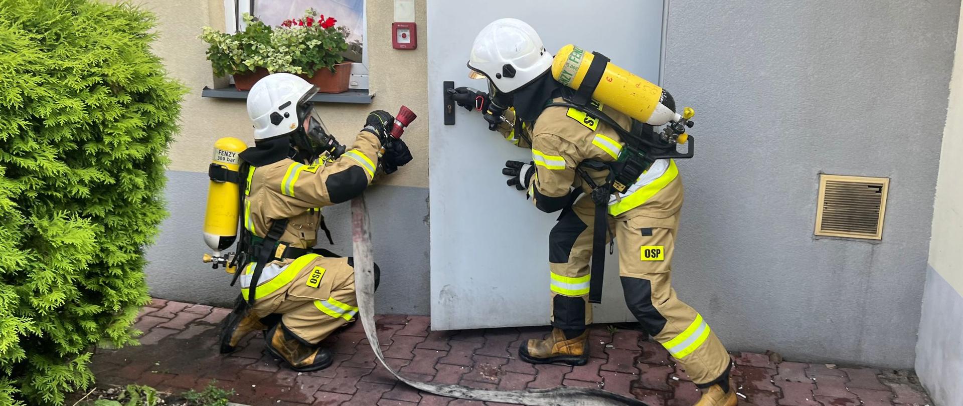 2 strażaków OSP ubranych w aparaty ochrorny dróg oddechowych, ubrania specjalne, biały hełm . jeden strażak otwiera drzwi do pomieszczenia, a drugi z wężem i prądownicą przed otwartymi drzwiami.
