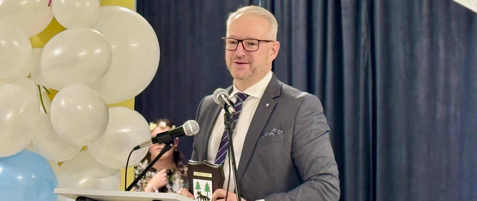 mężczyzna w szarym garniturze przemawia przez mikrofon na mównicy, za nim widać balony i kawałek transparentu 