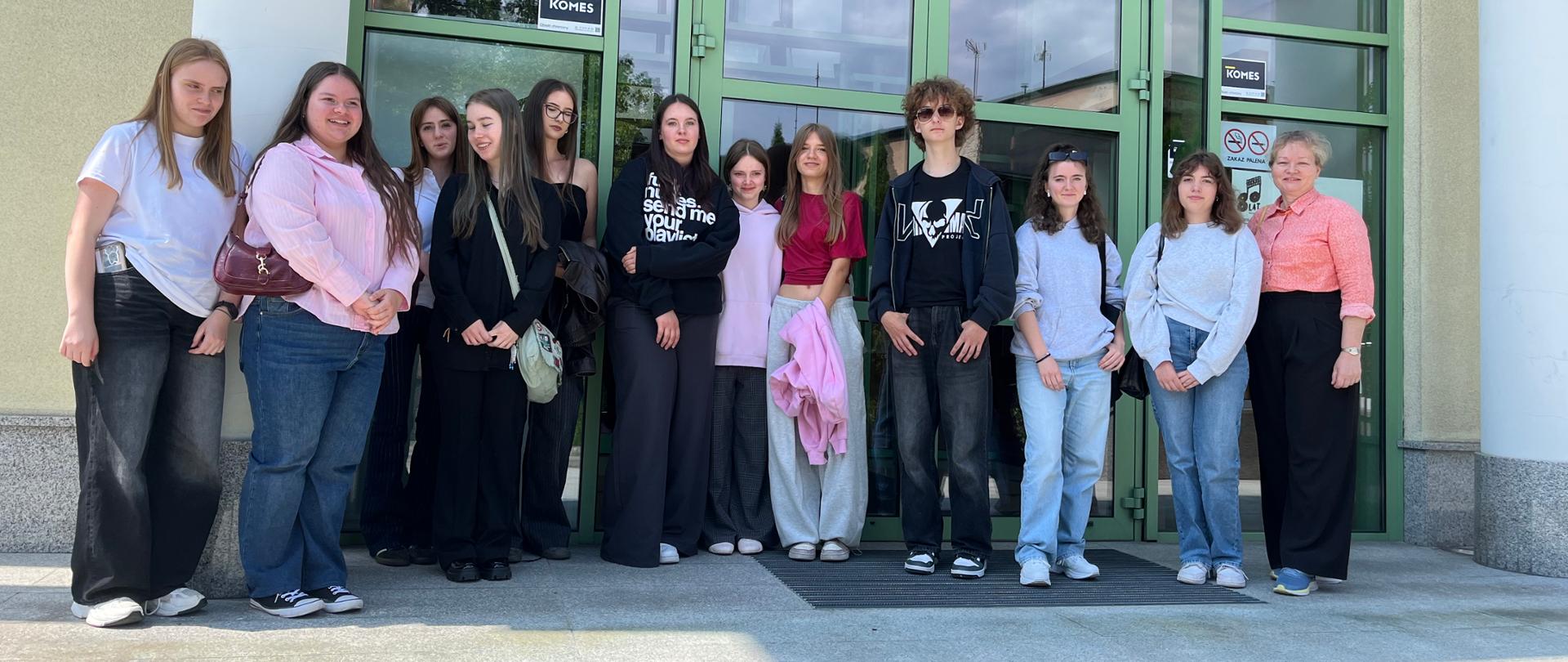 Grupa trzynastu osób stoi w szeregu przed wejściem do budynku z dużymi, zielonymi, przeszklonymi drzwiami. Wszyscy pozują do zdjęcia, ubrani swobodnie, w tle widać jasną elewację, odbicia chmur w szybach oraz naklejki informacyjne na drzwiach.