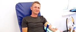 Na zdjęciu widoczny mężczyzna w czarnej koszulce i spodniach dżinsowych siedzi na fotelu koloru niebieskiego i oddaje krew w ramach akcji krwiodawstwa w budynku Komendy Powiatowej Państwowej Straży Pożarnej w Gorlicach
