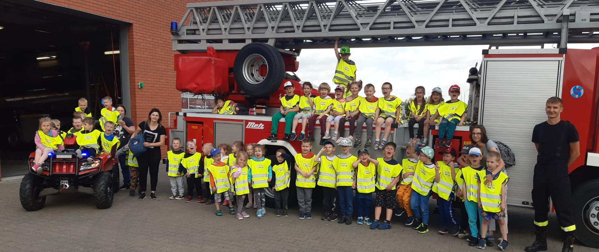 Przedszkolaki przed drabiną pożarniczą i na quadzie, z opiekunkami i strażakiem.