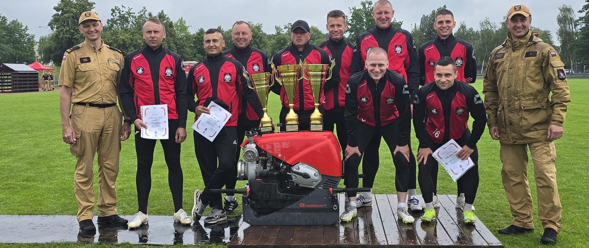 Zdjęcie grupowe na otwartym stadionie trawiastym przedstawia dziesięciu mężczyzn, w tym strażaków w umundurowaniu oraz zawodników w sportowych strojach strażackich, pozujących po zakończeniu zawodów.