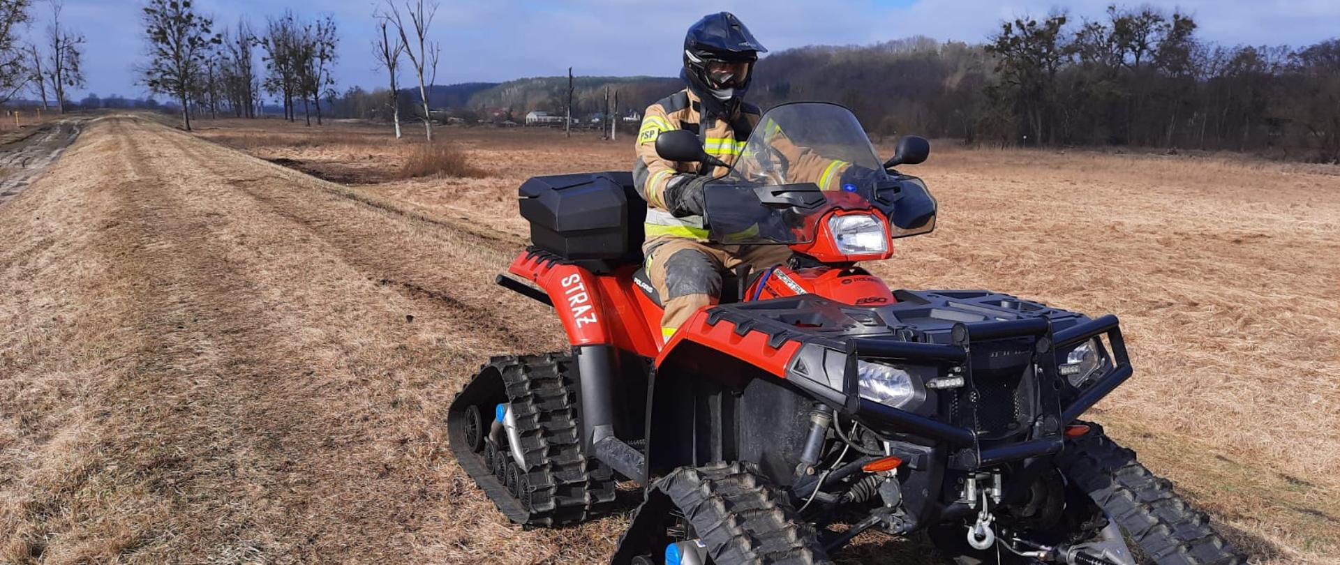 Fotografia przedstawia widok wzdłuż korony wału przeciwpowodziowego. Na pierwszym planie na quadzie z napędem gąsienicowym ustawionym czołem do fotografującego siedzi strażak w ubraniu specjalnym, z założonym kaskiem i goglami. W tle widoczne międzywale, porośnięte trawą.