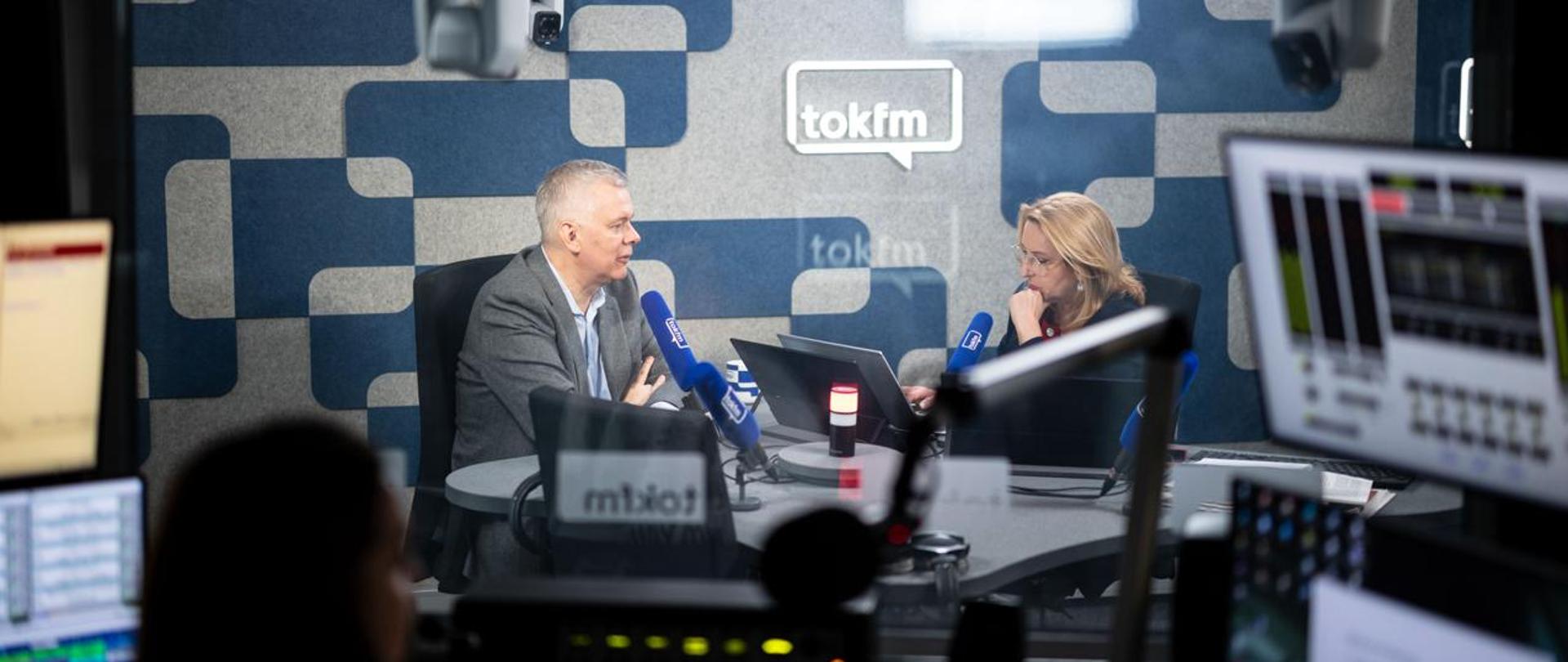 Na zdjęciu wnętrze studia radiowego. Przy stole, na czarnych krzesłach siedzą - minister Tomasz Siemoniak (po lewej) oraz redaktor Dominika Wielowieyska (po prawej). Na stole m.in.: cztery niebieskie mikrofony z napisem tok fm, biało-niebieski kubek, monitor oraz czerwona lampka. W tle ścianka w szaro-niebieskie wzory z neonem tokfm. U góry zdjęcia widać fragmenty dwóch białych kamer. Na pierwszym planie siedząca tyłem wydawczyni, mikrofon, mikser radiowy. Po prawej i lewej stronie zdjęcia widać monitory.