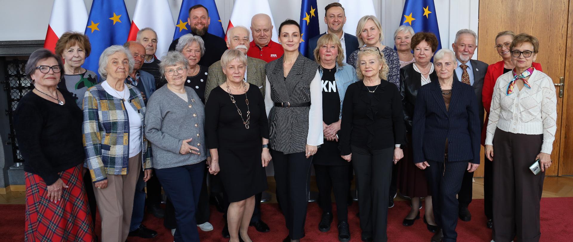 grupa osób, głownie w starszym wieku, pozuje do zdjęcia na tle polskich flag