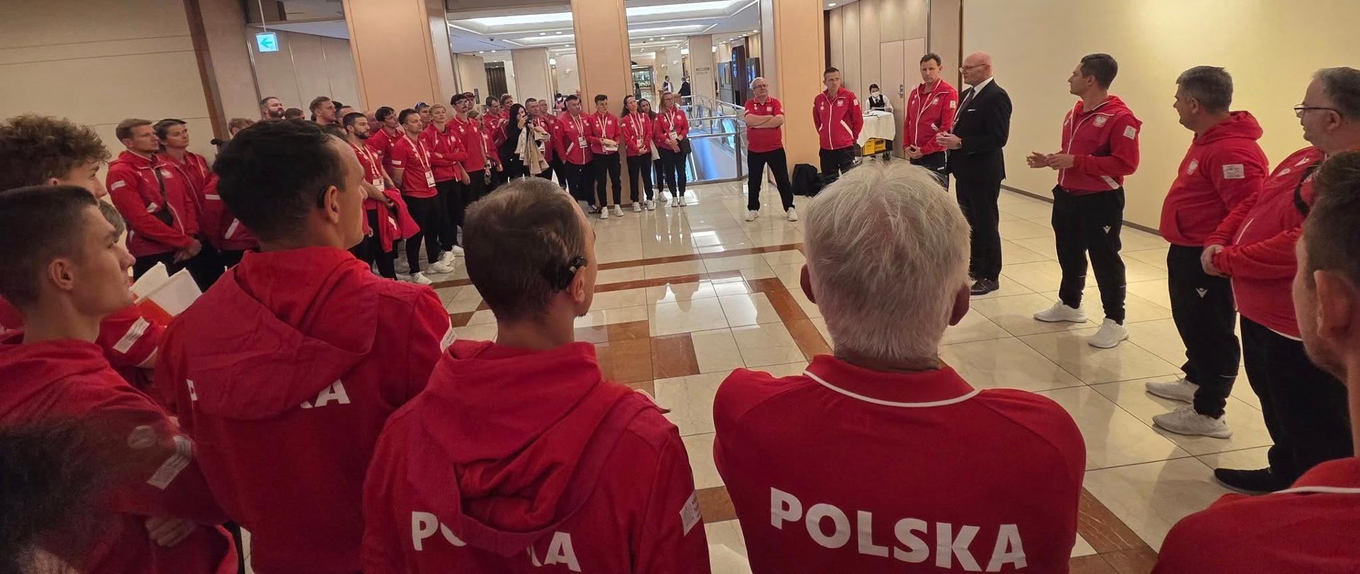 Zdjęcie przedstawiające spotkanie podsekretarza stanu Ireneusza Nalazka z reprezentacją polski na XXV Letnie Igrzyska Głuchych Tokio 2025. Minister stoi w hallu hotelowym otoczony dużą grupą osób w czerwonych bluzach z napisem "Polska" na plecach i czarnych spodniach.