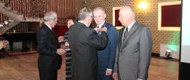 Na zdjęciu widzimy dwóch mężczyzn w dojrzałym wieku oraz kobietę stojących w szeregu. Do szeregu podchodzi kolejnych dwóch mężczyzn i przypinają sobą w szeregu odznaczenia. Wszyscy ubrani wyjściowo garnitury, koszule, krawaty. Dalej w tle widzimy wystrój sali, pozłacane ściany, kinkiety. 