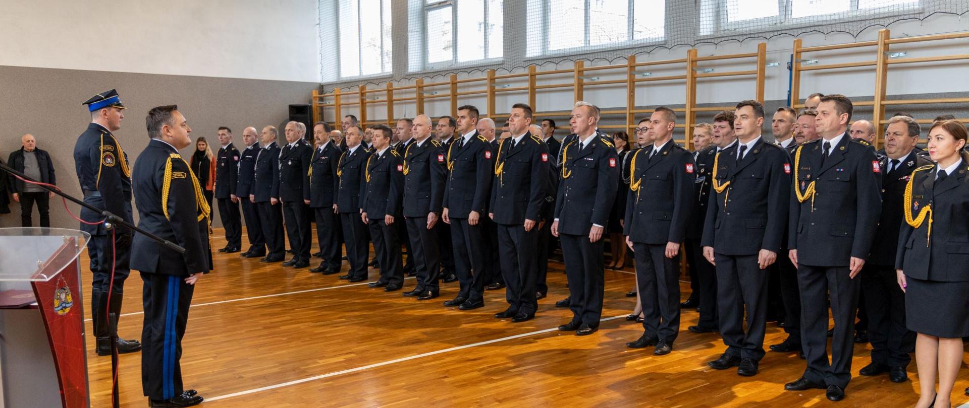 Strażacy Państwowej Straży Pożarnej oraz ochotniczych straży pożarnych stoją w czterech rzędach podczas uroczystości. Uroczystość na sali gimnastycznej Komendy Wojewódzkiej Państwowej Straży Pożarnej w Opolu. Strażacy podczas powitania Opolskiego Komendanta Wojewódzkiego PSP nadbryg. Krzysztofa Kędryka.