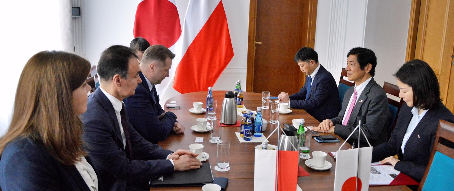Zdjęcie z boku, przy drewnianym stole siedzi grupa elegancko ubranych osób, przed nimi na stole małe flagi Polski i Japonii, za nimi białe ściany, w nich drewniane drzwi, pod ścianą duże flagi, z lewej zasłonięte białą firanką okno.
