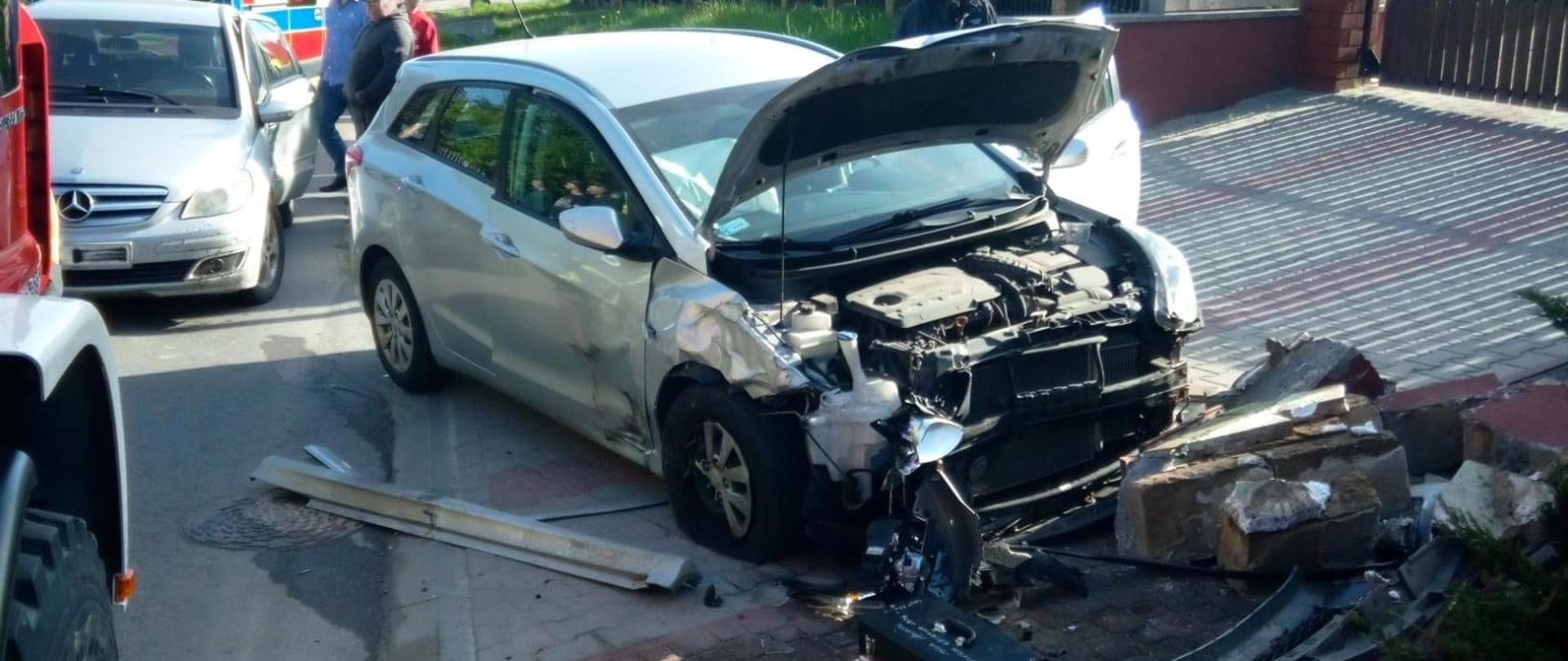 Zdjęcie przedstawia stojący na wjeździe na posesję samochód osobowy, drugi z uszkodzonych aut stoi na jezdni, lekko z tyłu. W głębi widać ambulans.
