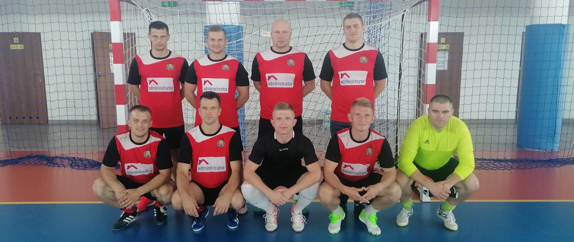 Zdjęcie przedstawia reprezentację Komendy Miejskiej Państwowej Straży Pożarnej w Radomiu podczas eliminacji strefowych strażaków w piłkę nożną. Strażacy ustawieni są na tle bramki w dwóch rzędach – w pierwszym rzędzie stoją, w drugim znajdują się w pozycji przykucniętej. Reprezentanci ubrani są w różnobarwne stroje piłkarskie (koszulki, spodenki). 