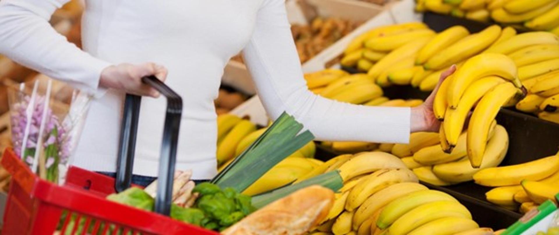 Kobieta w sklepie, trzyma w prawej ręce czerwony koszyk z zakupami spożywczymi, stoi przy półce z bananami, w lewej ręce trzyma kiść żółtych bananów.