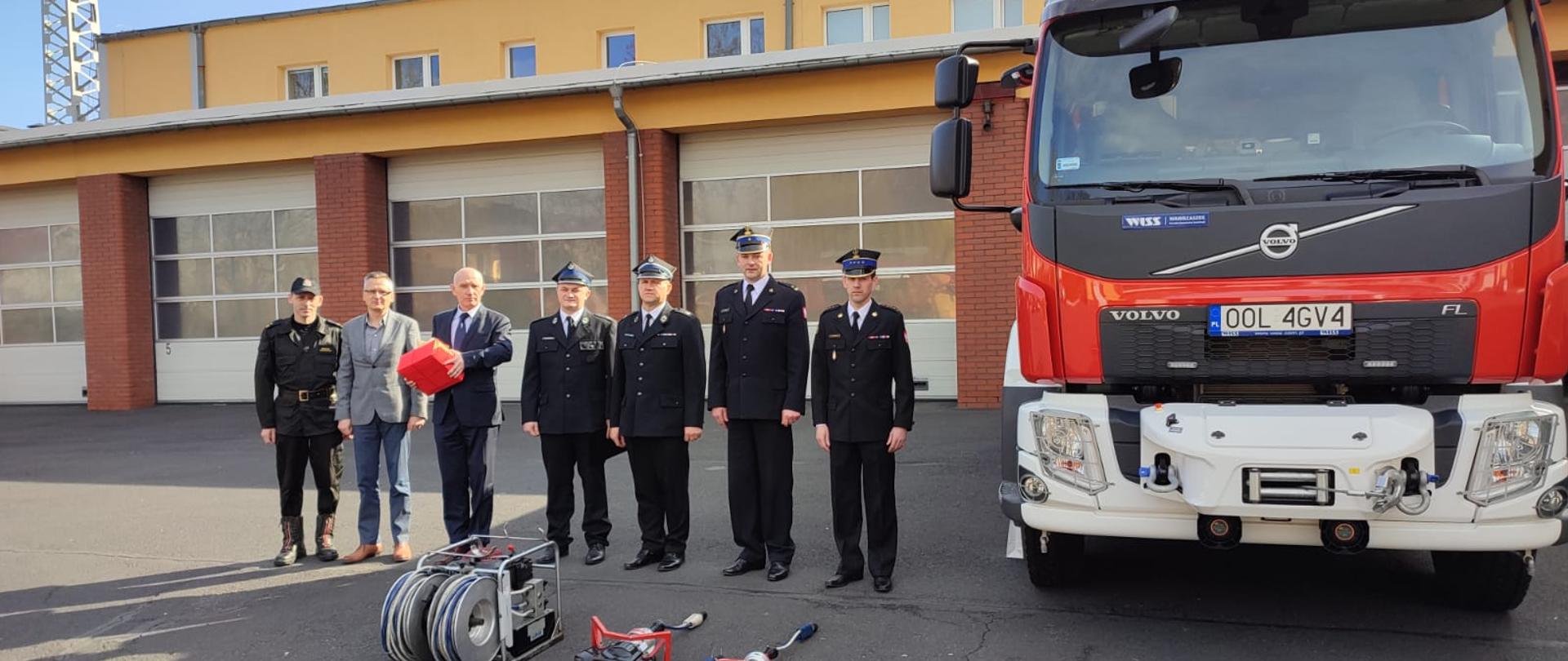 Przed garażami remizy strażackiej stoją strażacy, osoby cywilne, sprzęt hydrauliczny oraz samochód pożarniczy.