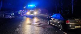 Na zdjęciu widoczny stojący po środku samochód strażacki z włączonymi sygnałami błyskowymi. Po prawej stronie zdjęcia od tyłu samochód osobowy marki Opel koloru ciemnego. Po lewej stronie zdjęcia w przydrożnym rowie samochód osobowy marki Opel koloru jasnego. Przy samochodzie strażackim stoją strażacy i policjant. Zdjęcie ciemnej tonacji, zrobione po zmroku. Widoczna mokra od deszczu jezdnia i odbite w niej światła.