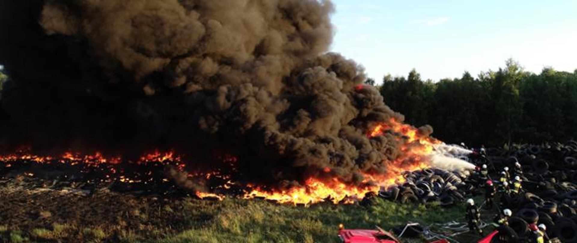 Widok pożaru składowiska opon na dużej powierzchni, spowitej gęstym, czarnym dymem. Na czole pożaru strażacy prowadzący działania gaśnicze pianą.