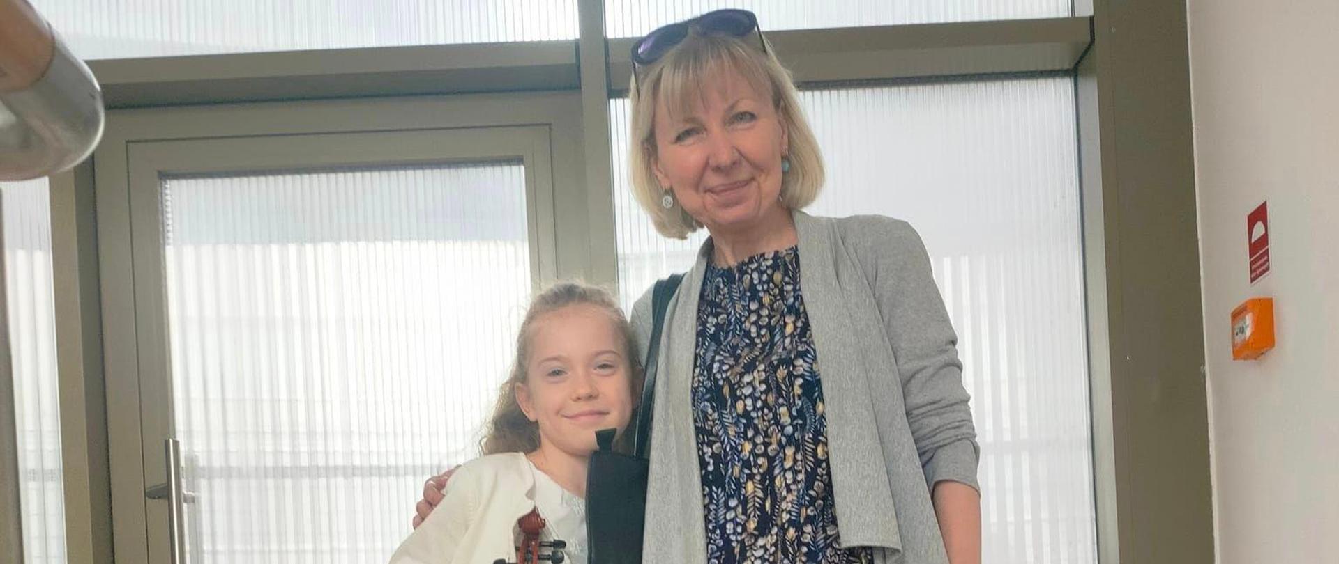 Uczennica z nauczycielką, w rękach trzyma skrzypce i smyczek, stoją na tle drzwi