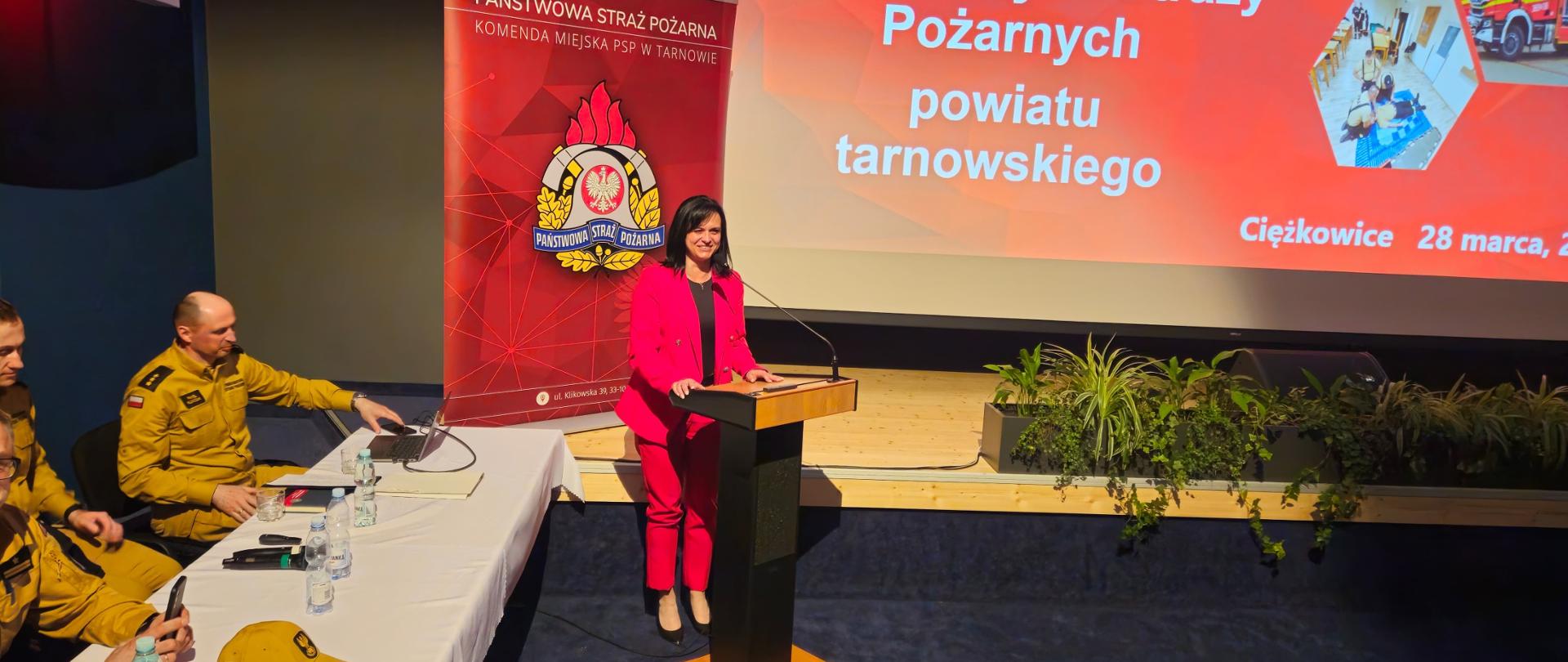 Na ekranie wyświetlona jest prezentacja, na scenie widoczne kwiaty, po prawej stronie przemawiający goście, oraz stół przy którym siedzą strażacy ubrani w piaskowe mundury